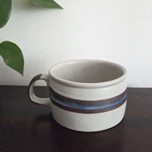 May include: A ceramic mug with a cream-colored base and a brown handle. The mug features two brown stripes with a blue stripe in between. The mug is sitting on a dark brown surface.