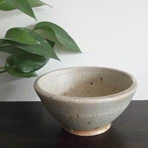 May include: A round ceramic bowl with a speckled, off-white and light gray glaze. The bowl features a wide, open rim and a tapered base with visible brown clay tones near the bottom, showcasing a textured, hand-crafted appearance throughout.