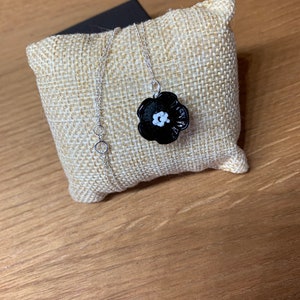 May include: A silver chain necklace with a black flower pendant with a white center. The necklace is displayed on a beige burlap pillow, set against a wooden surface.