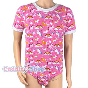 May include: Pink short-sleeved bodysuit with a white trim and a pattern of unicorns and rainbows. The bodysuit has the text "Cuddlz Shop" on the front.