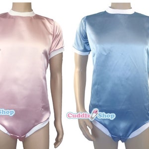 Peut inclure: Deux bodies en satin, un rose et un bleu, avec des bordures blanches sur le col et les manches. Les bodies ont des manches courtes et une fermeture à boutons-pression. Le logo Cuddlz Shop est visible en bas à gauche.