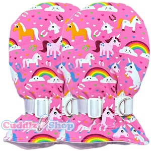 Könnte beinhalten: Rosa Fäustlinge mit einem sich wiederholenden Muster aus Einhörnern, Regenbogen und Hufeisen. Die Fäustlinge haben verstellbare weiße Riemen mit silbernen Schnallen. Der Text "Cuddlz Shop" ist unten aufgedruckt.