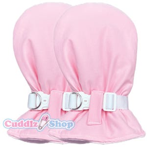Pode incluir: Duas luvas rosa com tiras e fivelas brancas ajustáveis. As luvas são feitas de um tecido macio e têm uma forma arredondada. As tiras prendem-se à volta do pulso. O logótipo Cuddlz Shop é visível.