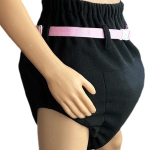 Puede incluir: Pañal de tela para adultos de color negro con una cinta ajustable de color rosa y una hebilla blanca. El pañal tiene una forma contorneada y una cintura elástica fruncida.