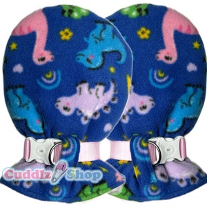 May include: A pair of blue fleece baby mittens with a dinosaur print. The mittens have pink straps with a buckle closure. The text "Cuddlz Shop" is visible in the bottom center of the image.