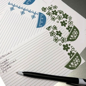 May include: A white lined notepad with a handwritten grocery list that reads "Groceries - bread - milk - blueberries - yogurt". A black pen is lying on the notepad. The notepad has a decorative border with blue and green floral designs.