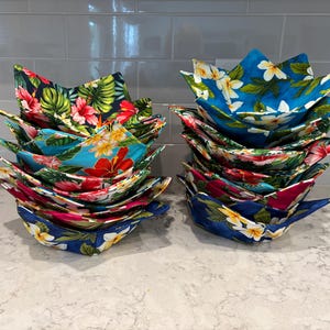 Peut inclure: Plusieurs piles de maniques pour bols en tissu aux motifs floraux vibrants. Les maniques présentent une variété de couleurs, dont le bleu, le rouge, le rose et le vert, avec des motifs de fleurs et de feuilles tropicales. Ces maniques sont conçues pour les bols chauds.