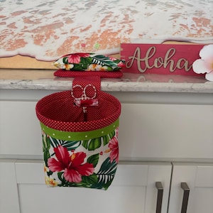 May include: A red and white polka dot fabric hanging basket with a green polka dot trim and a floral pattern of red hibiscus flowers and green leaves. A matching pin cushion and scissors are inside the basket. The word "Aloha" is on a wooden sign.