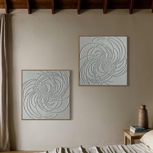 May include: Two framed abstract art prints with a white background and a swirling pattern of small white dots. The prints are hanging on a light beige wall.