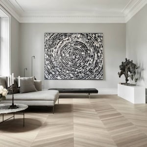 May include: A modern living room featuring a large abstract artwork with a circular pattern in shades of gray and white. The room includes a gray sofa, a black bench, and a sculpture on a white pedestal. The wooden floor has a chevron pattern.