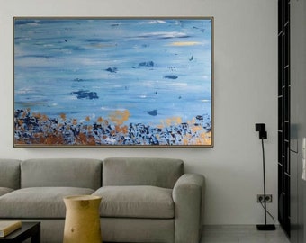 May include: Abstract painting with blue and gold hues. The painting features a textured, layered style with brushstrokes and splatters of paint. The painting is framed and hanging on a white wall in a living room setting.