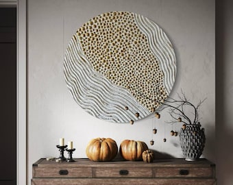 May include: A white and gold round wall decor with a textured surface and a pattern of small circles. The decor is hanging on a white wall above a wooden table with two pumpkins and a vase with a branch.
