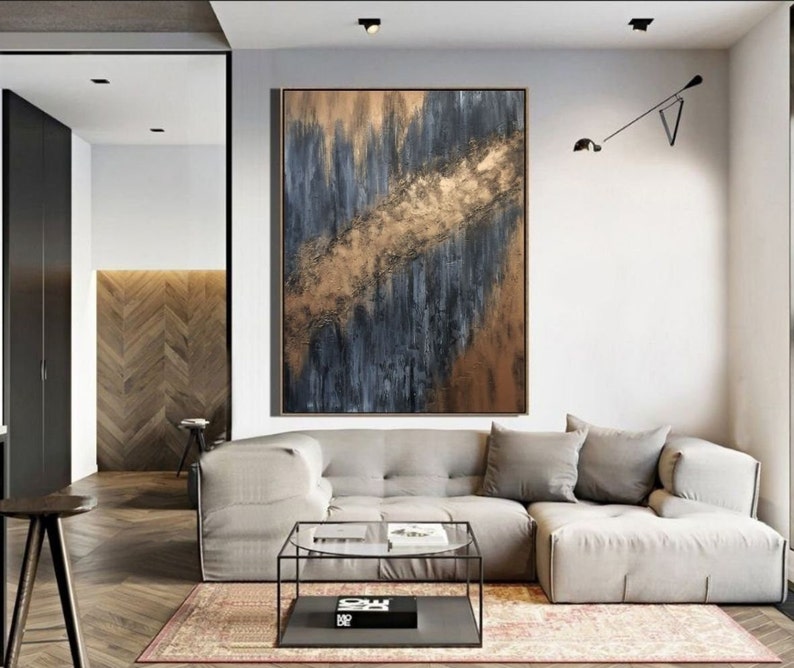 May include: Abstract painting with gold and blue hues on a canvas. The painting is hanging on a wall in a living room with a couch and coffee table.