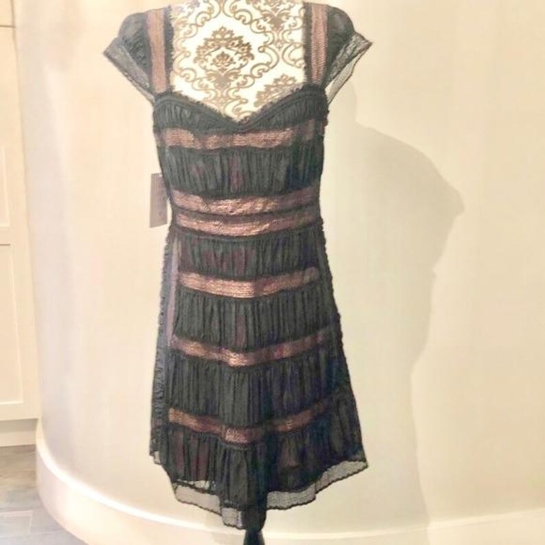 May include: Black knee-length dress with a sweetheart neckline and cap sleeves. The dress features horizontal bands of a copper-colored material, creating a layered effect. The dress is displayed on a mannequin.