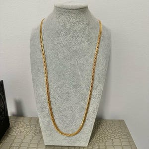 May include: A gold-colored chain necklace displayed on a gray mannequin. The chain has a curb link design and is draped in a U-shape. The necklace is a simple, elegant piece of jewelry.