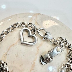 K&ouml;nnte beinhalten: Ein silberfarbenes Armband mit einem herzf&ouml;rmigen Anh&auml;nger und einem Karabinerverschluss. Das Armband hat ein Kettenglied-Design und ein kleines Herzdetail am Verschluss. Der Herzanh&auml;nger ist ein offenes Herz.