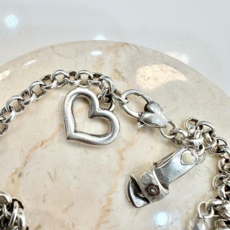 K&ouml;nnte beinhalten: Ein silberfarbenes Armband mit einem herzf&ouml;rmigen Anh&auml;nger und einem Karabinerverschluss. Das Armband hat ein Kettenglied-Design und ein kleines Herzdetail am Verschluss. Der Herzanh&auml;nger ist ein offenes Herz.