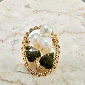 May include: A gold-toned brooch with an oval frame, featuring three white pearls and two dark green jade leaves. The brooch has a floral design, with the pearls arranged above the leaves.