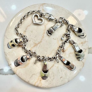 K&ouml;nnte beinhalten: Silbernes Charm-Armband mit mehreren Miniatur-Schuh-Anh&auml;ngern in verschiedenen Farben, darunter Rosa, Gelb und Gr&uuml;n. Das Armband enth&auml;lt einen herzf&ouml;rmigen Anh&auml;nger und kleine Kristallakzente. Das Armband wird auf einer hellen, strukturierten Oberfl&auml;che pr&auml;sentiert.