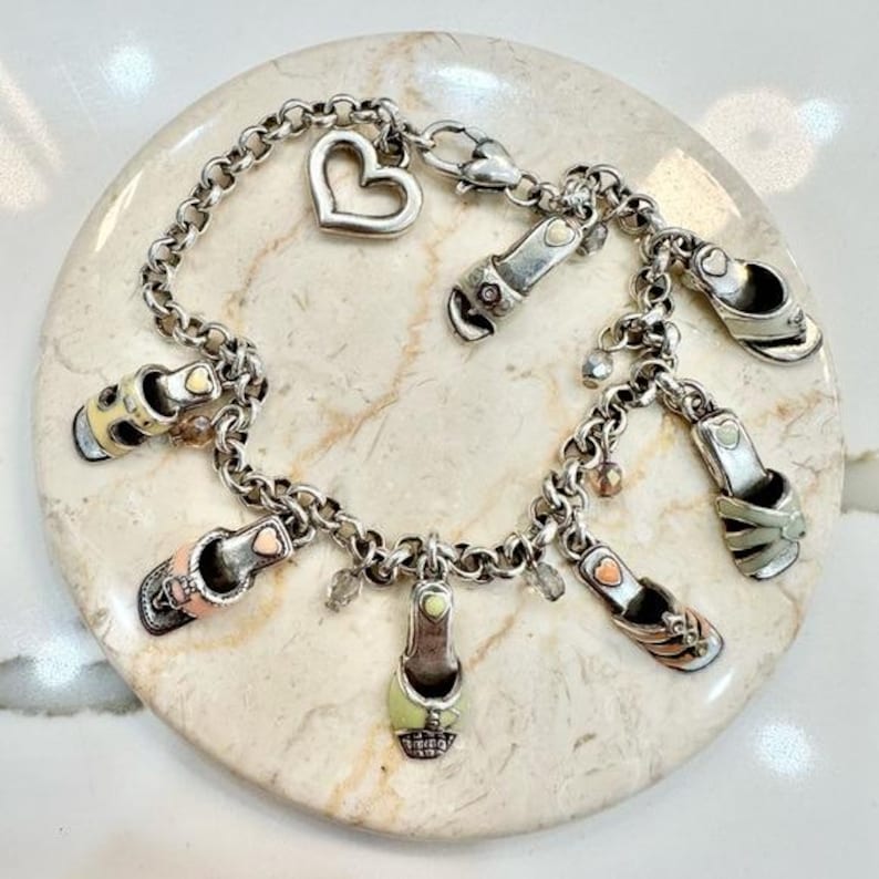 K&ouml;nnte beinhalten: Silbernes Charm-Armband mit mehreren Miniatur-Schuh-Anh&auml;ngern in verschiedenen Farben, darunter Rosa, Gelb und Gr&uuml;n. Das Armband enth&auml;lt einen herzf&ouml;rmigen Anh&auml;nger und kleine Kristallakzente. Das Armband wird auf einer hellen, strukturierten Oberfl&auml;che pr&auml;sentiert.