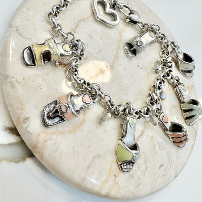 K&ouml;nnte beinhalten: Ein silbernes Charm-Armband mit verschiedenen Schuh-Anh&auml;ngern in Pastellfarben. Das Armband enth&auml;lt Charms mit gelben, rosa und gr&uuml;nen Akzenten sowie einen herzf&ouml;rmigen Charm. Das Armband wird auf einer hellen, strukturierten Oberfl&auml;che pr&auml;sentiert.