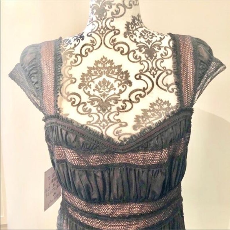 May include: A black dress with a sweetheart neckline and cap sleeves. The dress features a layered design with lace and ruffled accents. The straps and bodice have a pinkish-tan trim. The dress is displayed on a mannequin with a decorative pattern.