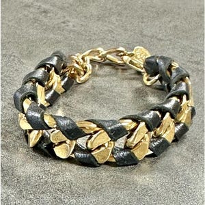 May include: A braided bracelet featuring alternating black leather and gold-toned metal links. The bracelet has a chain closure and a small gold charm. The jewelry is displayed on a gray surface.
