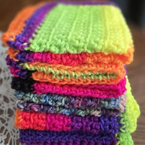 May include: A stack of colorful, handmade crochet squares. The squares are in vibrant shades of neon green, orange, pink, purple, and black. The stack is on a wooden surface, with a white lace doily visible in the background.