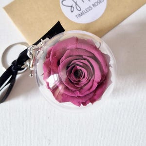 May include: A pink preserved rose encased in a clear glass dome with a black keychain attached. The dome is labeled "S&Fleur Timeless Roses".