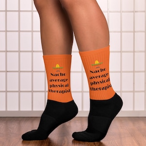 May include: A pair of crew socks featuring a bright orange upper section and a solid black lower section. The orange part displays a small graphic of a sombrero above the black text that reads "Nacho average physical therapist" in a bold font.