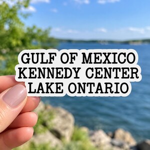 Op de afbeelding: Een witte, contourgesneden sticker met een zwarte rand en zwarte tekst die in drie afzonderlijke regels GULF OF MEXICO, KENNEDY CENTER en LAKE ONTARIO leest. De sticker heeft een golvende, onregelmatige rand die de vorm van de vette schreefloze letters volgt.