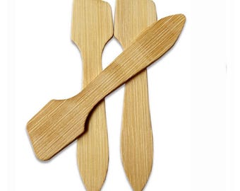 May include: Four small, light brown bamboo spatulas with a flat, angled head and a long, narrow handle. The spatulas are arranged on a white background, with one overlapping the others. The bamboo has a visible wood grain.