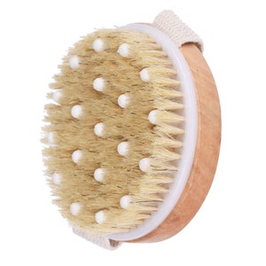 Puede incluir: Un cepillo corporal redondo de madera con cerdas naturales y protuberancias de plástico blanco. El cepillo tiene una correa de algodón blanco.