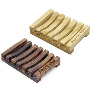 Può includere: Due portasapone in legno, uno marrone chiaro e uno marrone scuro. Entrambi hanno un design curvo e il testo "Engrave Logo" è inciso sul fondo.