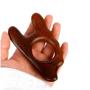 May include: A dark brown wooden massage tool with a smooth, polished finish. The object features an irregular, organic shape with multiple rounded protrusions and a large, circular opening in the centre, designed for ergonomic grip and use.