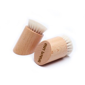 Può includere: Due spazzole per la pulizia del viso in legno con setole bianche. Lo spazzolino a destra ha le parole "ENGRAVE LOGO" incise sul lato.