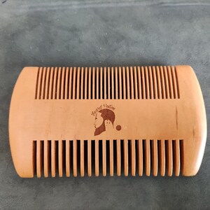 Könnte beinhalten: Ein Holzbartkamm mit brauner Oberfläche. Der Kamm hat eine abgerundete Form und trägt ein Logo mit dem Text "Beard Notion" und einem stilisierten Bild eines bärtigen Mannes.