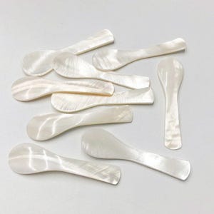 May include: A collection of small, pearlescent white spoons. Each spoon has a rounded bowl and a flat, rectangular handle. The spoons are arranged on a white surface, showcasing their iridescent sheen and delicate design. Ideal for serving caviar or other small portions.