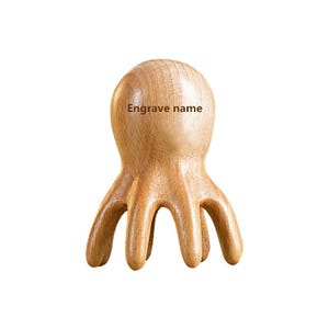 May include: A wooden octopus-shaped massage tool with a rounded head and eight curved tentacles. The text "Engrave name" is printed on the head of the octopus. The tool is made of light-coloured wood.