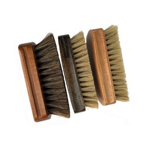 Puede incluir: Cuatro cepillos de zapatos de madera con cerdas naturales están dispuestos en diagonal sobre un fondo blanco. Los cepillos tienen diferentes tonos de madera marrón y colores de cerdas, de claro a oscuro.