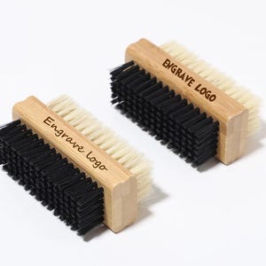 Könnte beinhalten: Zwei Nagelbürsten aus Holz mit natürlichen und schwarzen Borsten. Der Text "Engrave Logo" ist auf dem Holzgriff jeder Bürste aufgedruckt. Die Bürsten befinden sich auf einer weißen Oberfläche.