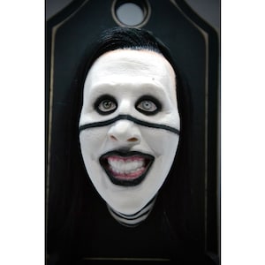 May include: A black and white painted face with a wide smile, the eyes are outlined in black with a black stripe across the nose. The face is framed by black hair and is set against a dark background.