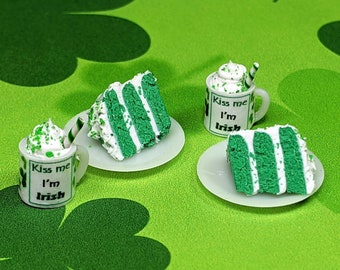 May include: Miniature St. Patrick's Day treats on a green clover-patterned surface. The image features two slices of green layered cake with white frosting, two mugs with whipped cream, and the text "Kiss me I'm Irish."