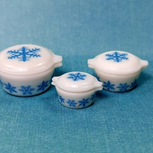 May include: Set of three white ceramic casserole dishes with blue snowflake designs. The dishes are nested, with the largest dish at the bottom and the smallest dish at the top.