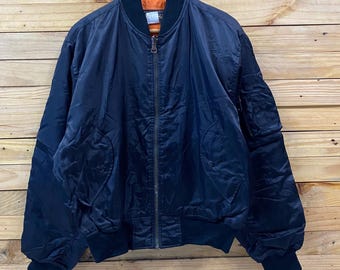 May include: A navy blue bomber jacket with a black collar, cuffs, and waistband. The jacket has a zipper closure and two front pockets. The interior lining is orange. The jacket is hanging on a wooden hanger against a wooden background.