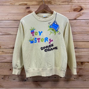 K&ouml;nnte beinhalten: Ein gelber, lang&auml;rmliger Pullover mit einem Toy Story-Motiv, das die Figuren aus dem Film zeigt. Das Motiv enth&auml;lt den Text "Toy Story" und "Space Crane".