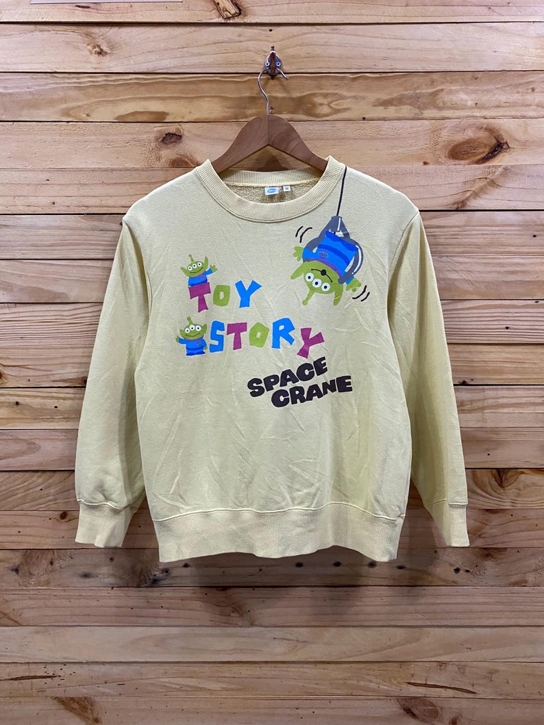 K&ouml;nnte beinhalten: Ein gelber, lang&auml;rmliger Pullover mit einem Toy Story-Motiv, das die Figuren aus dem Film zeigt. Das Motiv enth&auml;lt den Text "Toy Story" und "Space Crane".