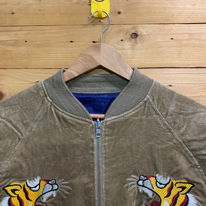 Pu&ograve; includere: Una giacca bomber in velluto a coste marrone con chiusura a zip e teste di tigre ricamate su entrambi i lati. La giacca ha una fodera blu.