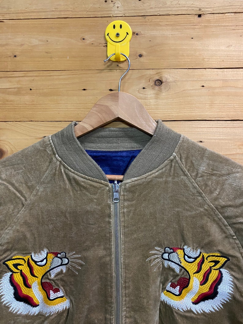 Pu&ograve; includere: Una giacca bomber in velluto a coste marrone con chiusura a zip e teste di tigre ricamate su entrambi i lati. La giacca ha una fodera blu.