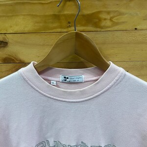 May include: A light pink Courr&egrave;ges Paris sweatshirt with a crew neck. The brand name is embroidered in silver thread across the chest. The sweatshirt is hanging on a wooden hanger against a wood-paneled background.
